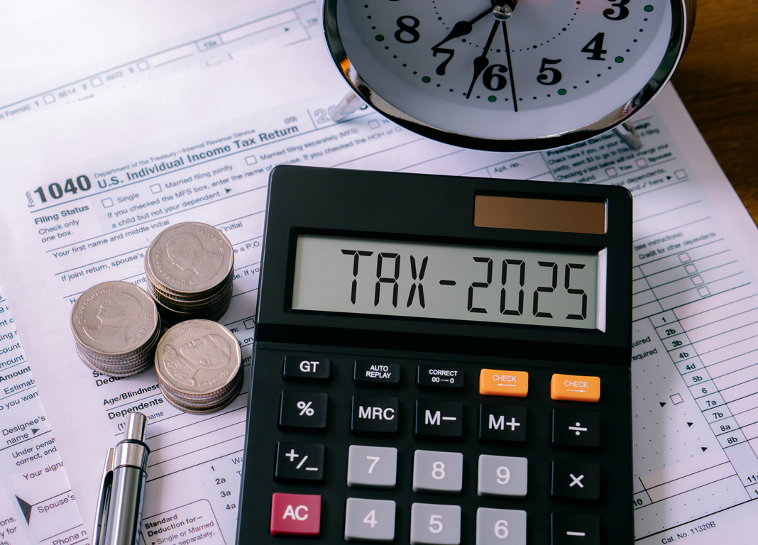 Stacks of coins and a pen on top of an IRS tax return, next to a calculator that reads "TAX-2025," beneath an alarm clock.